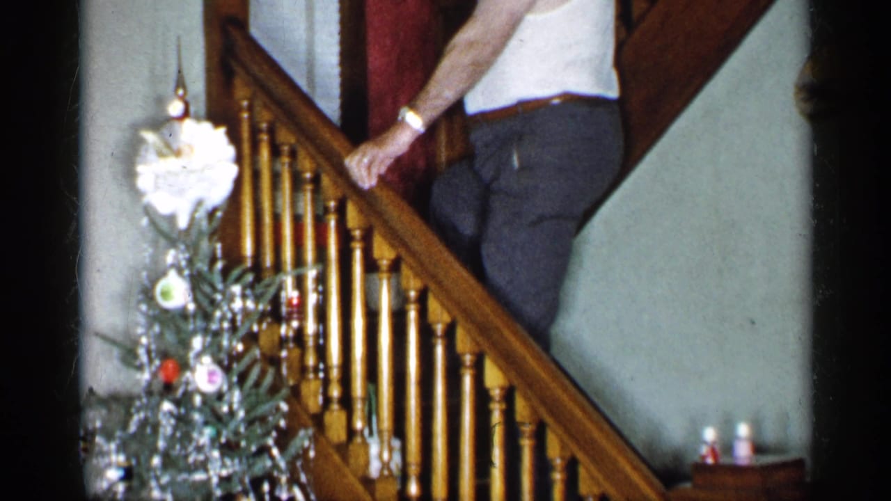 In a 1955 New York home-movie frame, an adult man climbs a wooden staircase near a tinsel-covered Christmas tree.