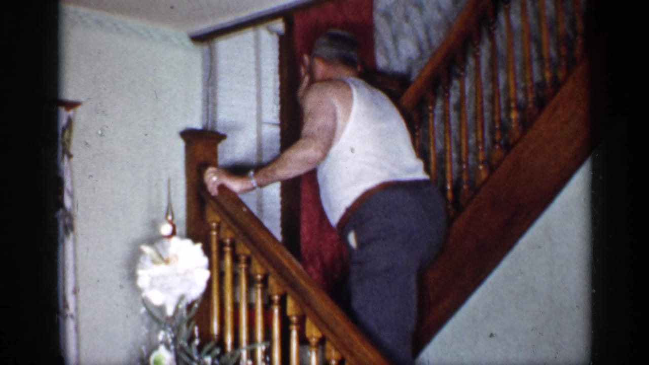Man in a white tank top climbs higher on the wooden stairs near the Christmas tree.