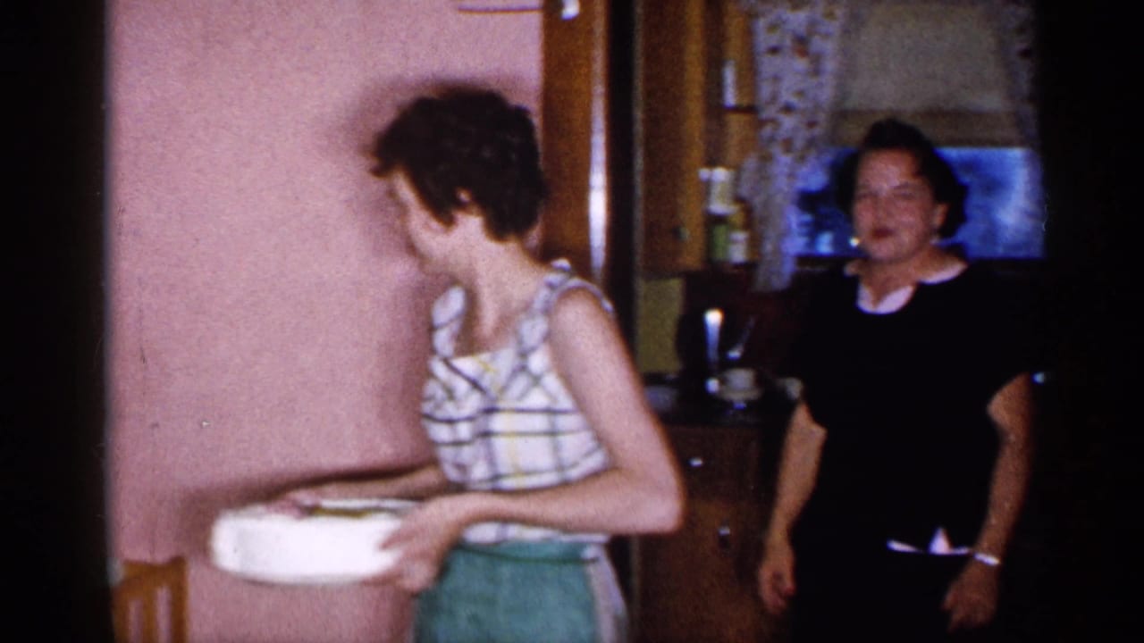 1955 New York home-movie frame of a woman in a plaid sleeveless top carrying a white cake or dish through a pink-walled interior.