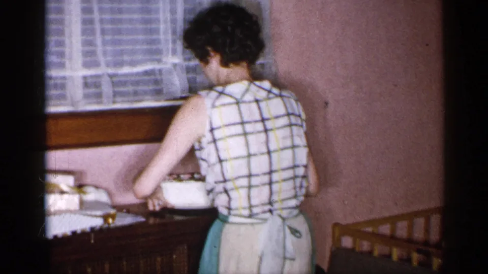 1955 New York home-movie frame showing a woman from behind setting a white cake or dish near a window and wooden furniture.