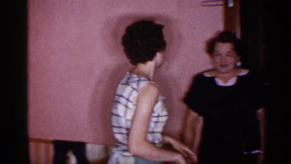 1955 New York home-movie frame with two women in a pink-walled nursery-like room beside a crib and wood doorway.