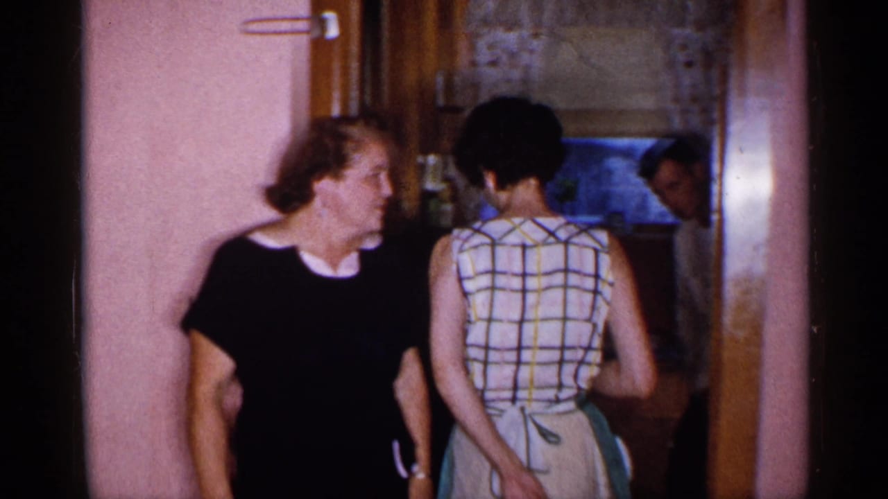 Two women and a man stand near a doorway into a small kitchen with pink walls.