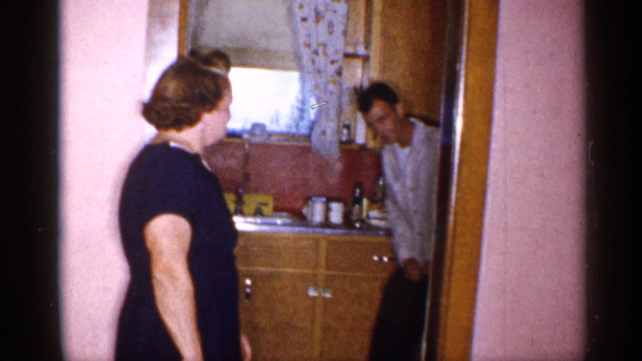 A woman stands beside the doorway of a small 1955 New York kitchen while a man leans in near wood cabinets and a sink area.