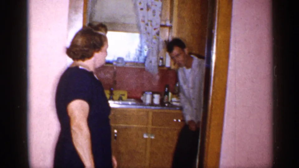A woman stands beside the doorway of a small 1955 New York kitchen while a man leans in near wood cabinets and a sink area.