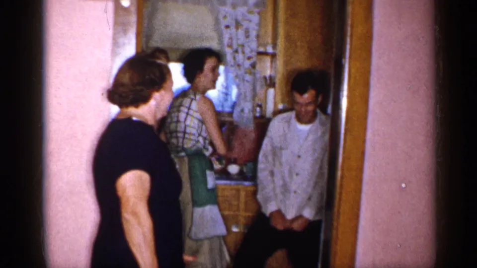 Two women stand near a pink-walled kitchen doorway while a man in a light shirt sits or crouches close to the cabinet area.