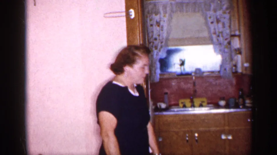 A woman in a black dress moves past a pink-walled doorway, with a small 1955 New York kitchen visible behind her.