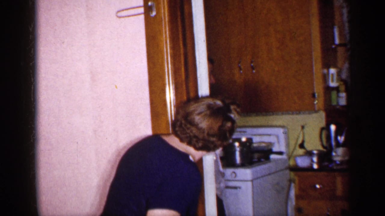Woman in a dark dress bends near a kitchen doorway with a stove and cabinets behind her.