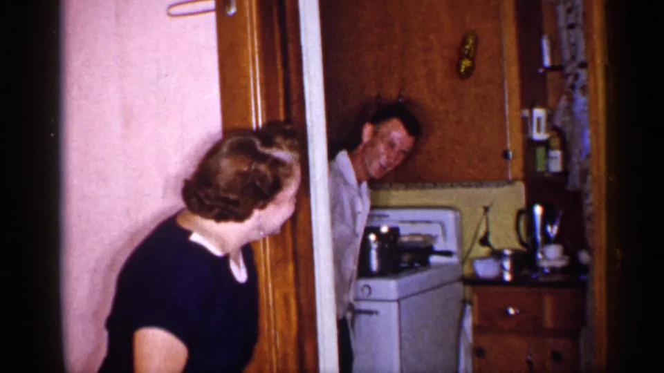 A woman turns back toward a man appearing from behind the doorway in a 1955 New York kitchen, with cabinets, stove, and cookware visible.
