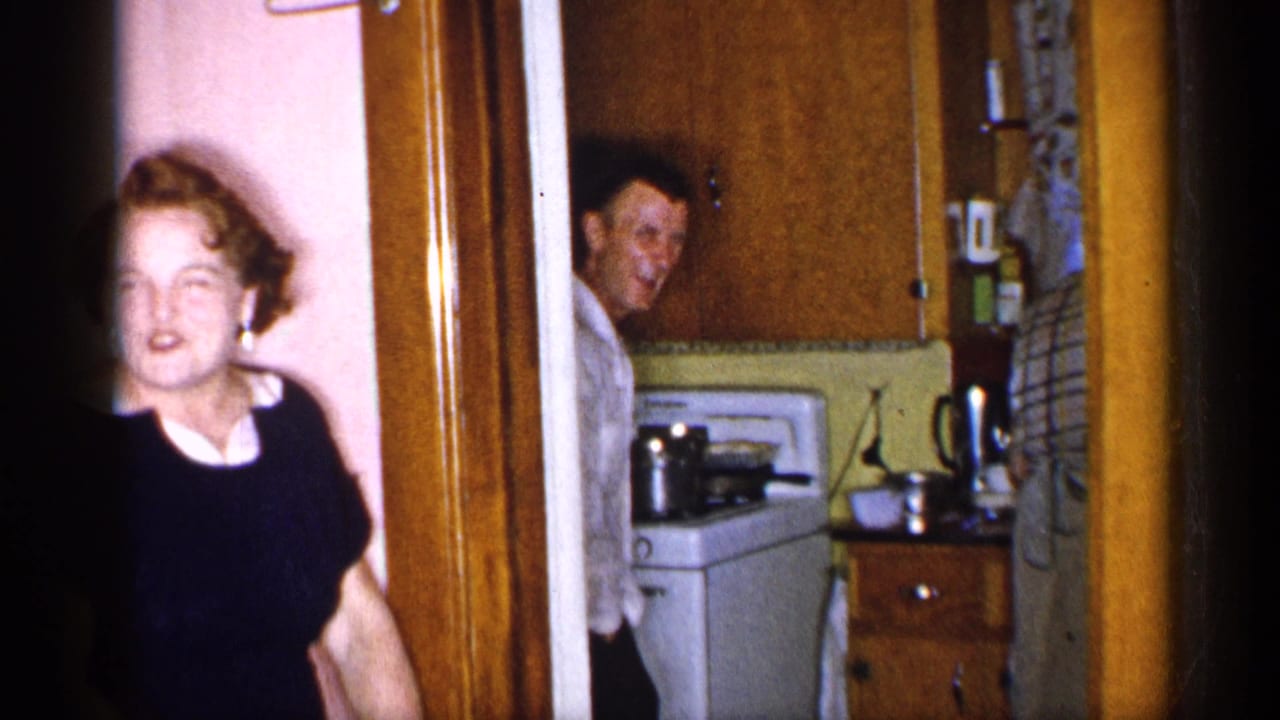 Woman in a dark dress near a doorway while a man looks from a small kitchen.