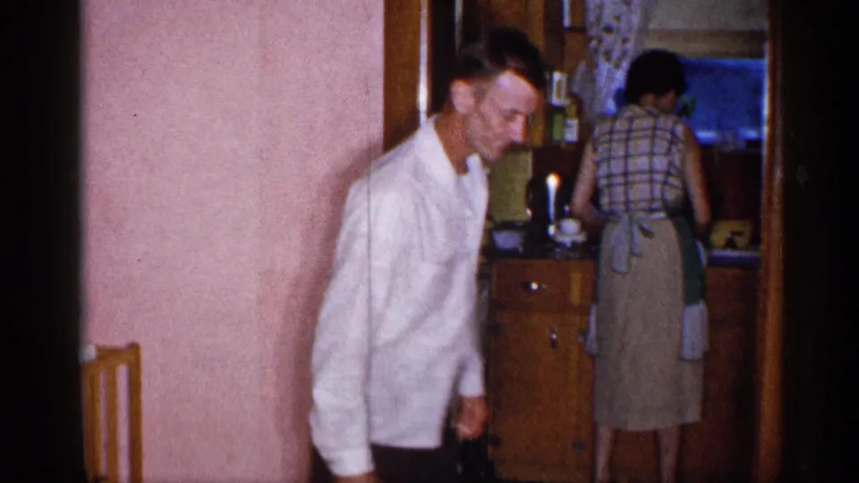 1955 New York home-movie frame showing a man by a doorway with an apron-clad woman at kitchen cabinets in the background.