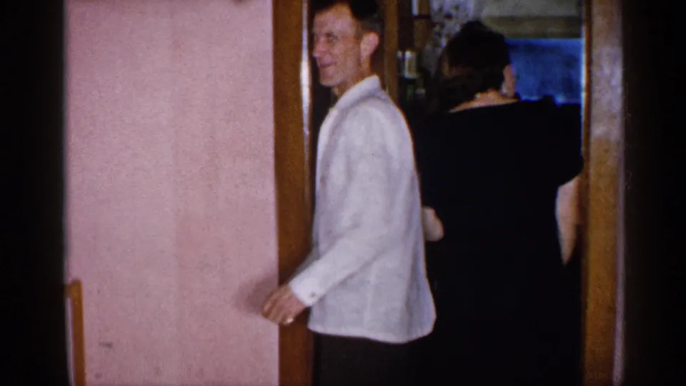 1955 New York home-movie frame with a smiling man in a white shirt and a woman in a dark dress inside a private home doorway.