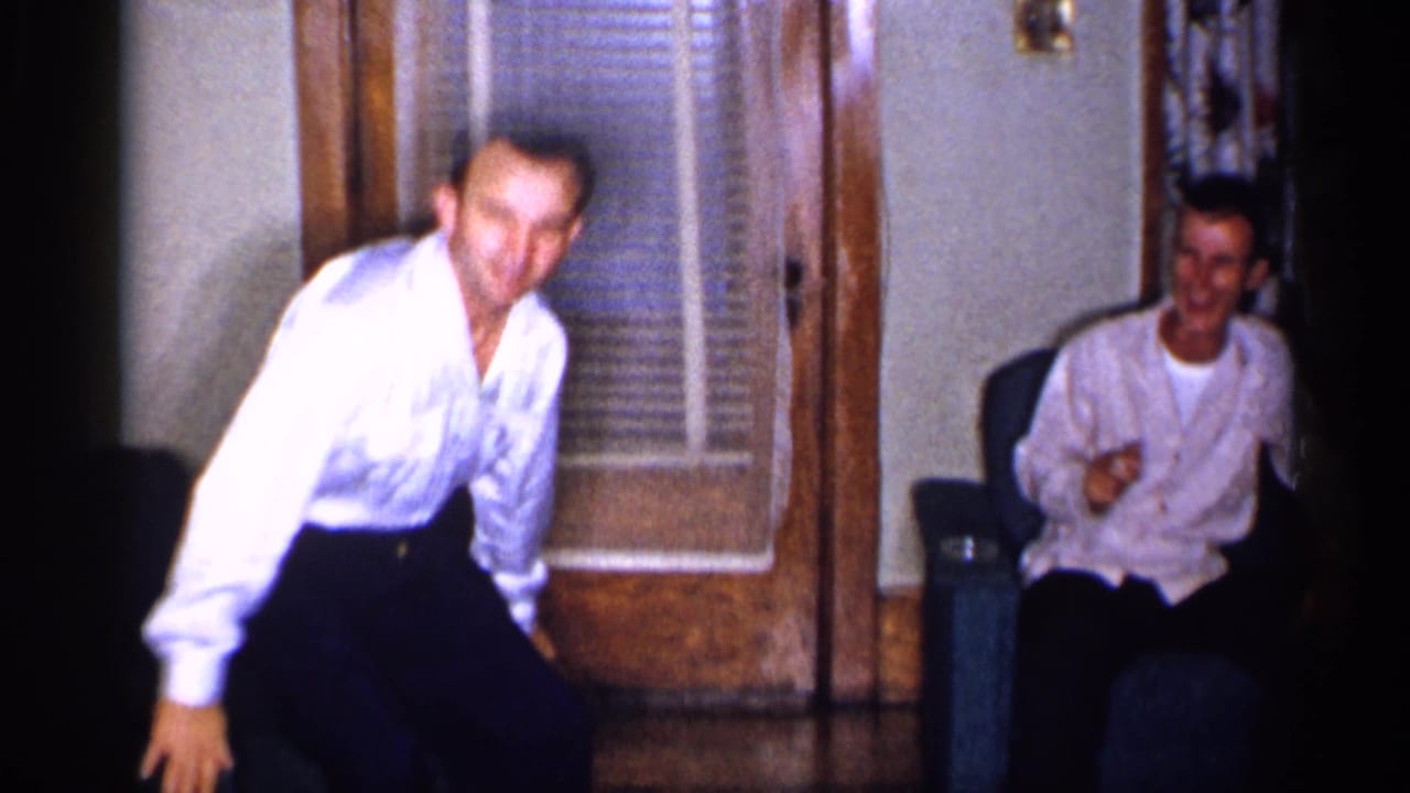 Adult man in a white shirt stands near a wood-framed door while another man sits on a green sofa.