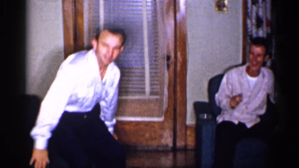 1955 New York home-movie frame with one adult man standing near a glass-paneled door and another seated in a living room.