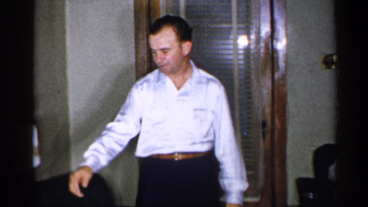 1955 New York interior frame showing an adult man standing in a white shirt near wood-framed windows and blinds.
