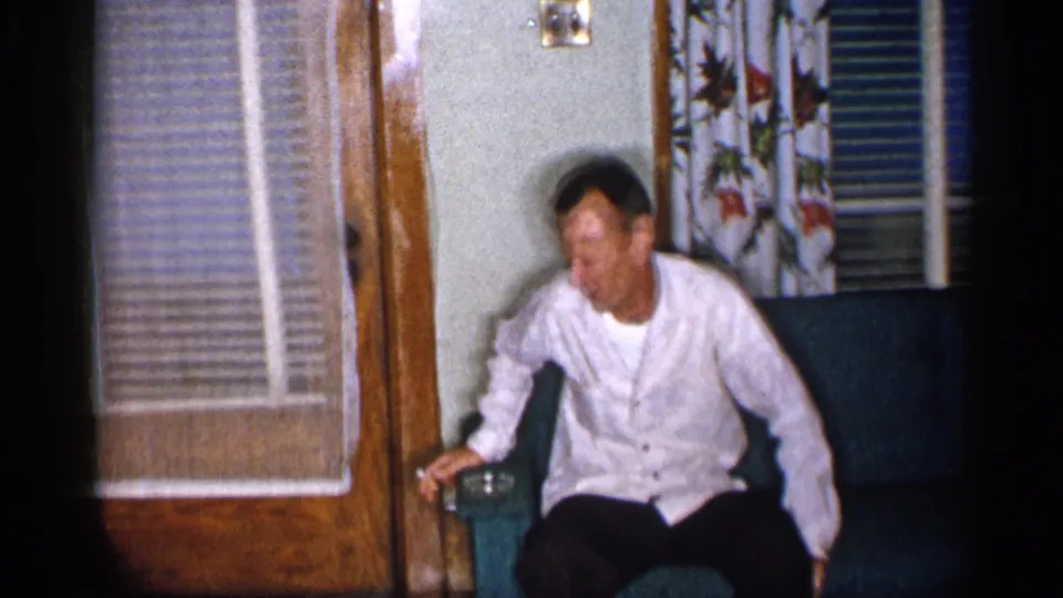 1955 New York home-movie frame of an adult man seated on a green sofa near floral curtains and a door with blinds.