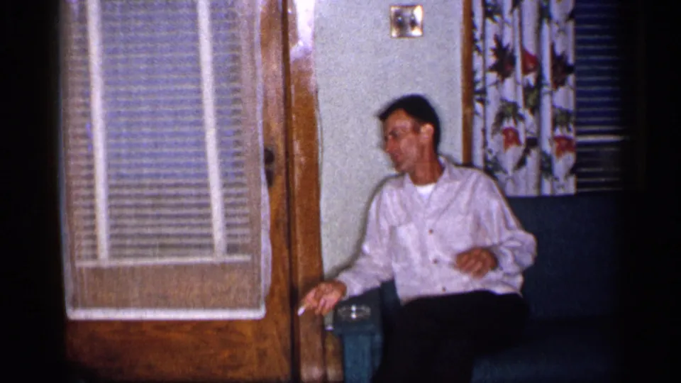 1955 New York living-room frame with an adult man seated on a green sofa, holding a small object near floral curtains.