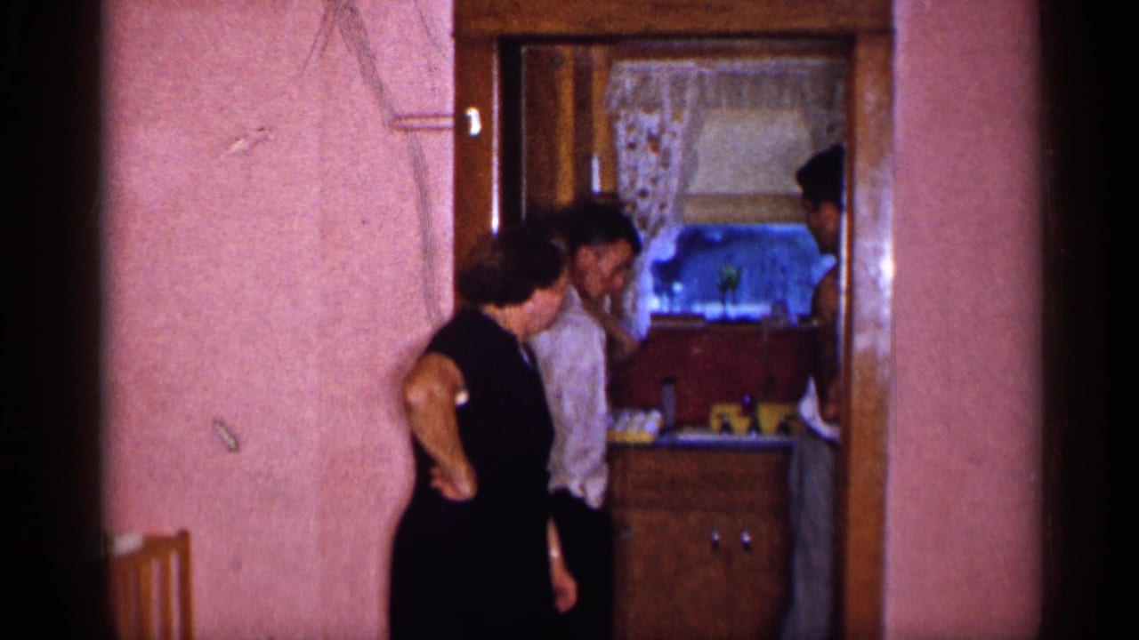 In the New York home-movie frame, adults stand at a doorway beside pink walls and wood trim, with a curtained room beyond.