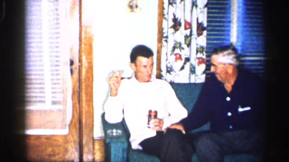 Two men sit together on a green couch in a 1955 New York home interior, with one holding a drink and pipe near floral curtains.