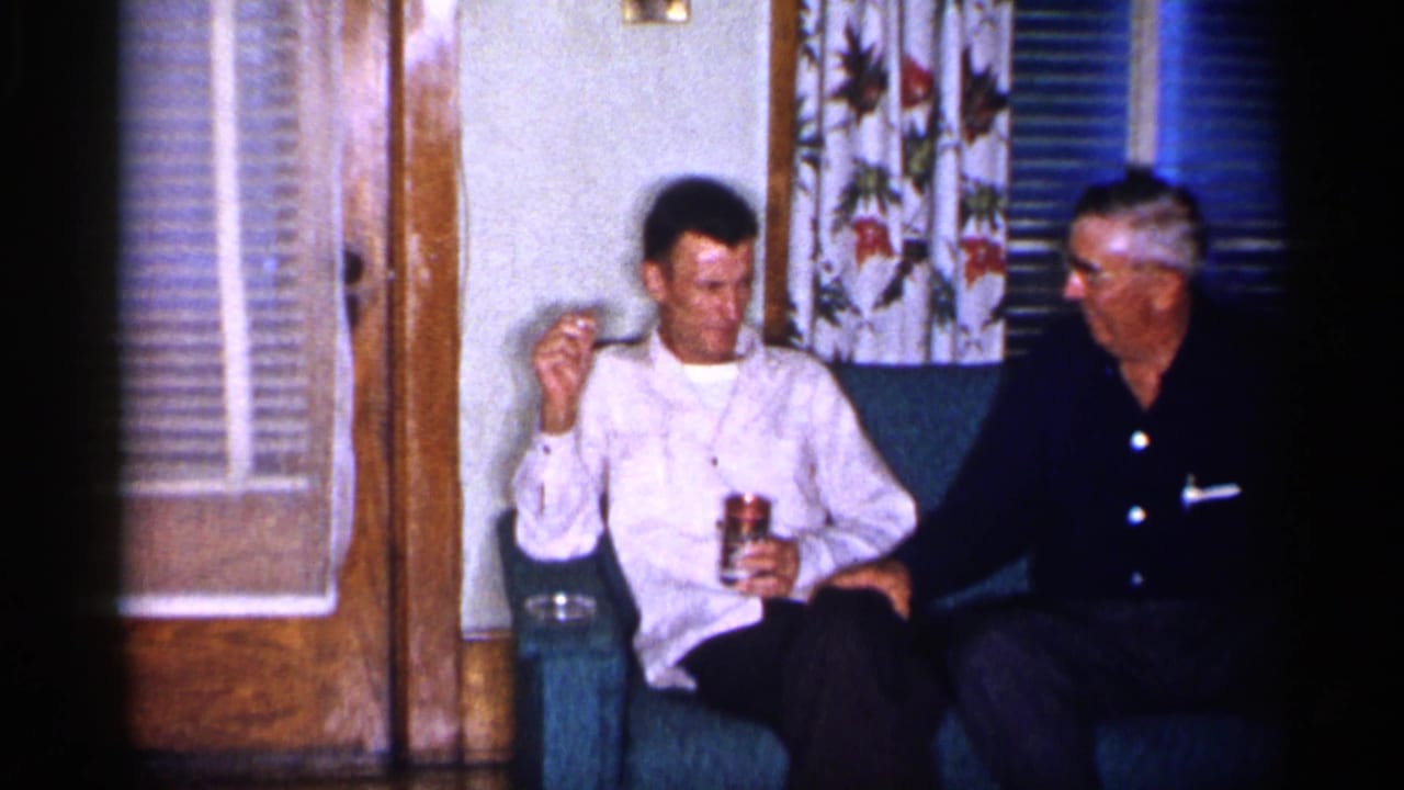 Two men sit on a teal couch, one holding a can and cigarette in a living room.
