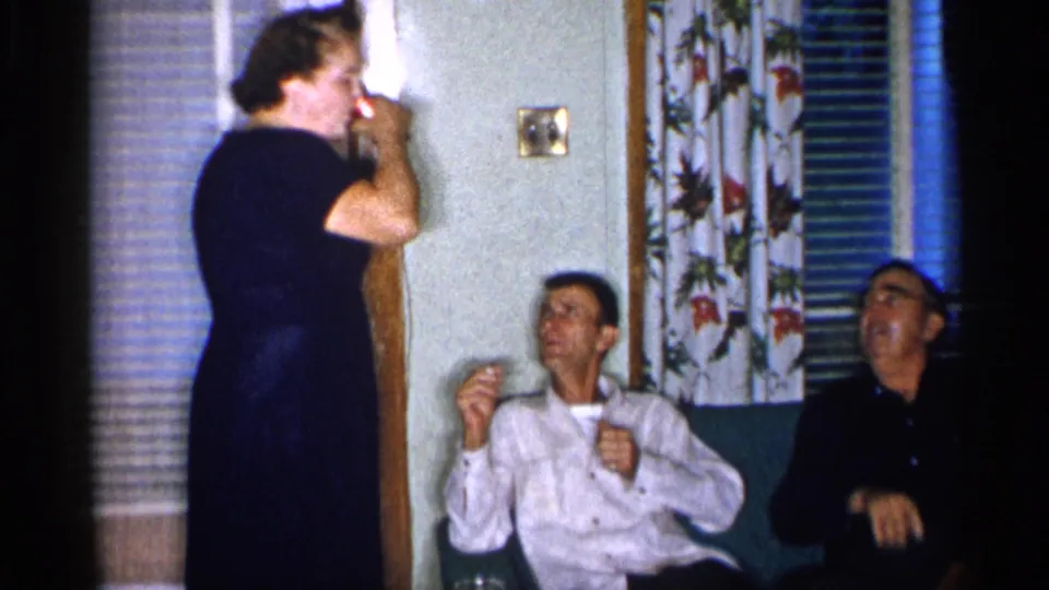 A woman in a dark dress stands in the 1955 New York living room holding a can while the two seated men look toward her.