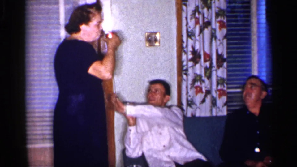 The woman stands near the couch with a can as the man in a white shirt gestures toward her and the other man sits beside him.