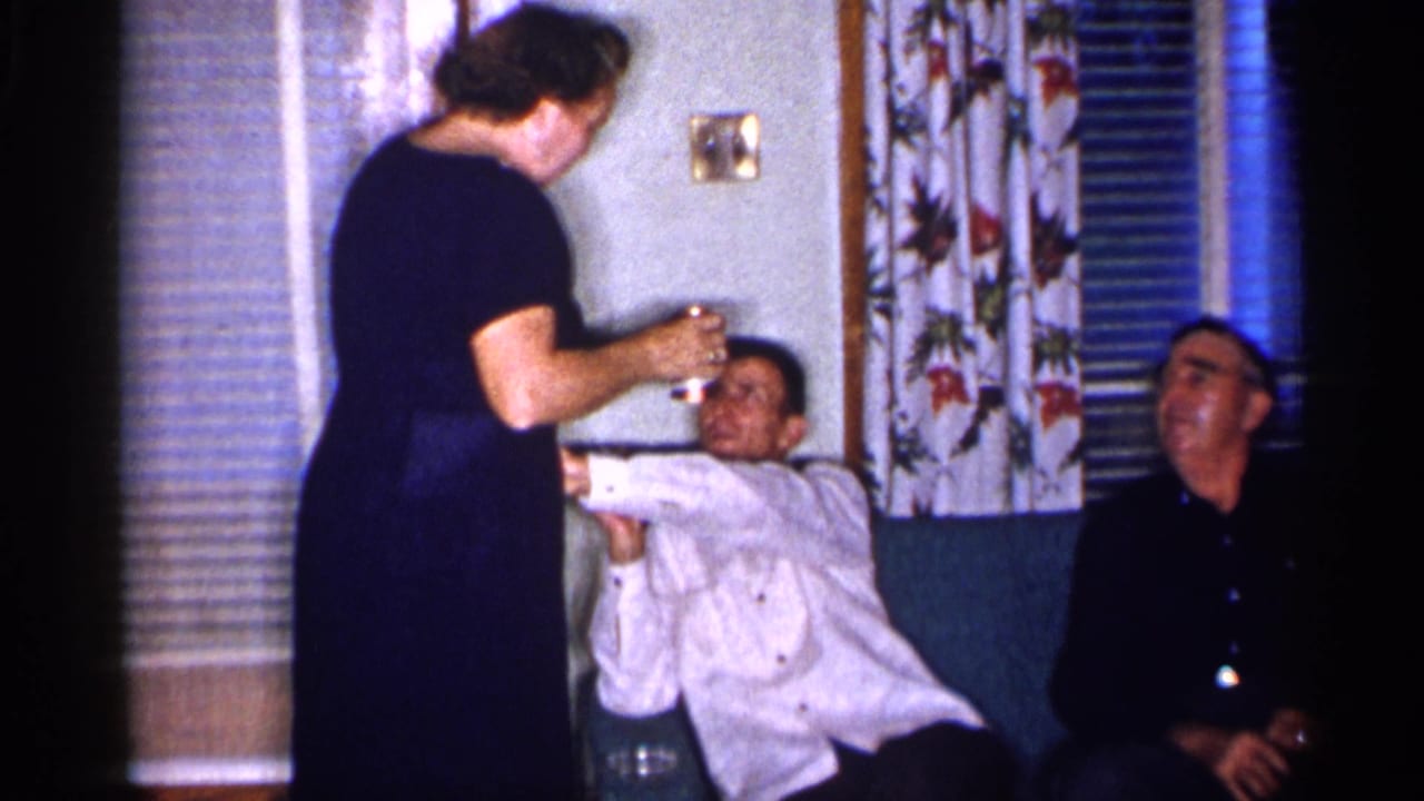 Woman in a dark dress stands near two seated men in a living room.