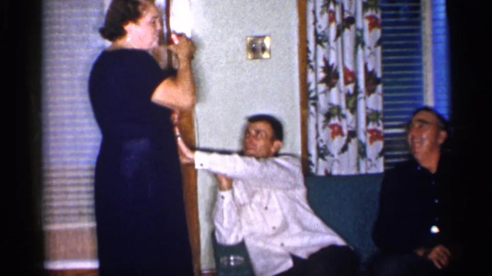 The woman raises her drink near her mouth while the seated man keeps one arm extended toward her in the living room.