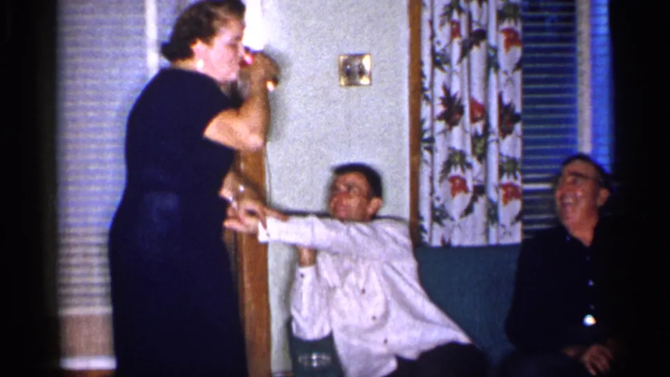 A woman in a dark dress leans forward holding a drink while the two seated men appear amused in the 1955 home-movie frame.