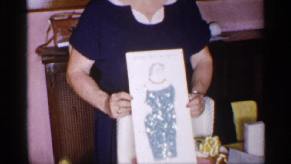 In a 1955 New York home-movie frame, a woman in a navy dress with a white collar holds up a framed illustration indoors.