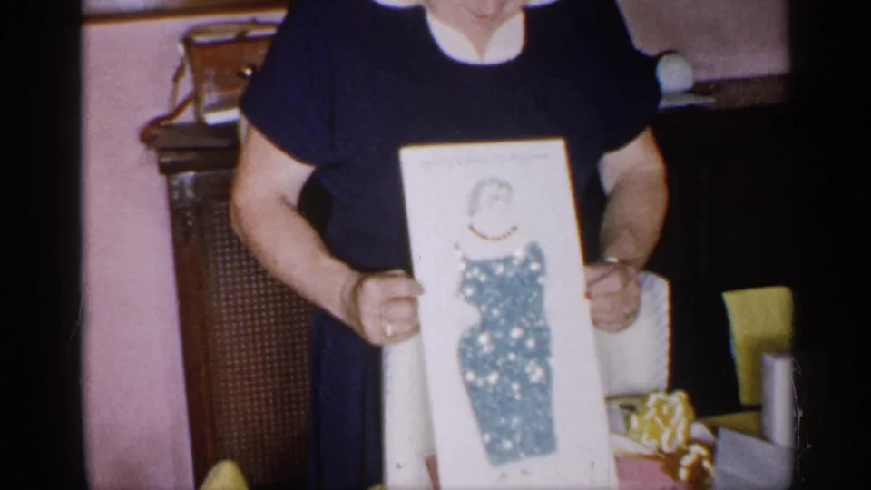 The woman continues displaying the framed blue-dress illustration in a private New York interior from the 1955 home movie.