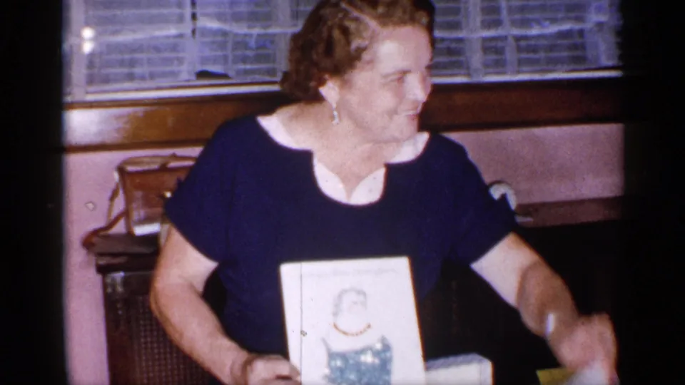 A smiling woman in a navy dress holds the framed illustration while seated or leaning in a mid-1950s New York living room.