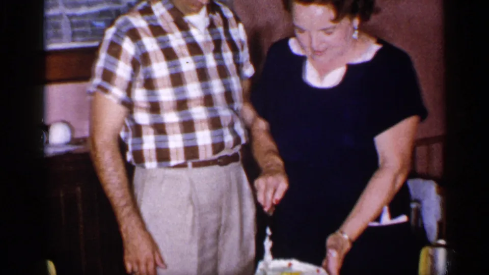 A man in a plaid shirt and the woman stand near a birthday cake in the 1955 New York home-movie celebration scene.