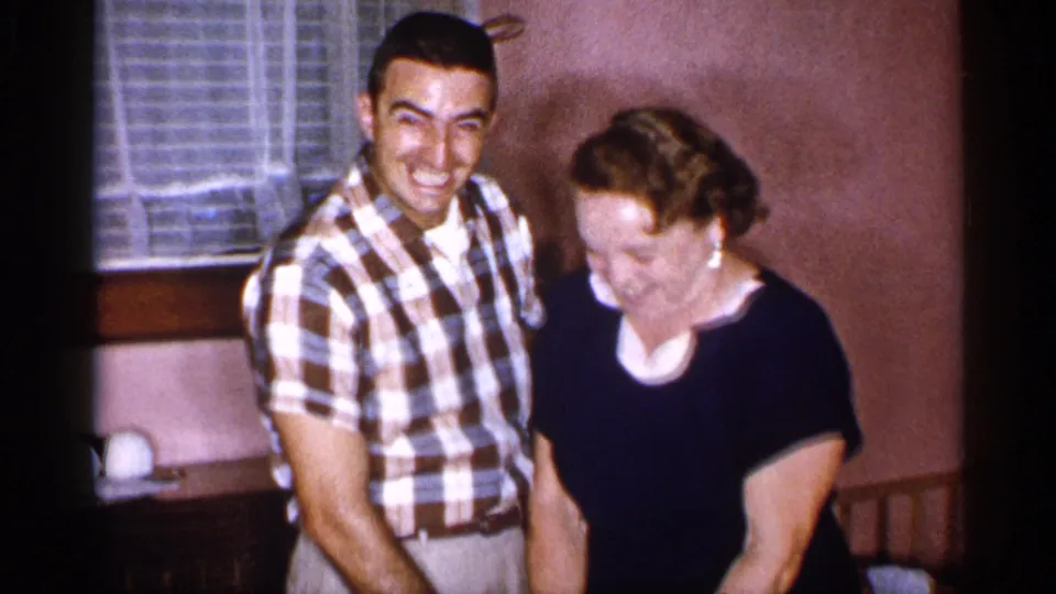 A smiling man in a plaid shirt stands beside a woman in a dark dress during the cake-cutting moment in the 1955 New York home movie.