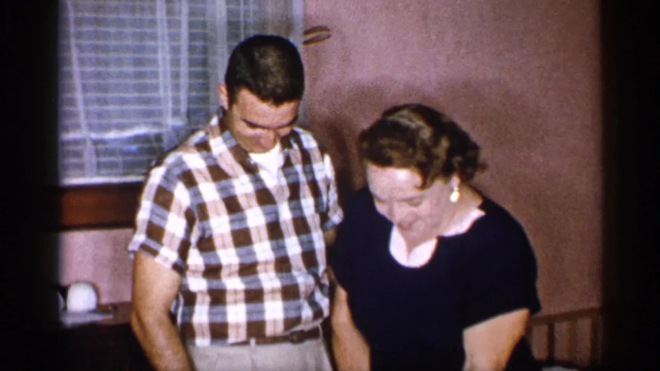 The couple looks down near the cake in a warm indoor 1955 New York home-movie frame, with window blinds and wall details behind them.