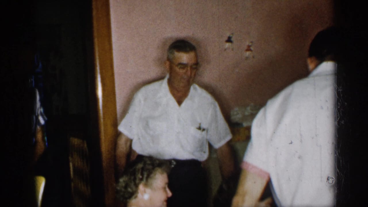 Adults gathered in a pink-walled dining room as a man in a white shirt stands near seated women.