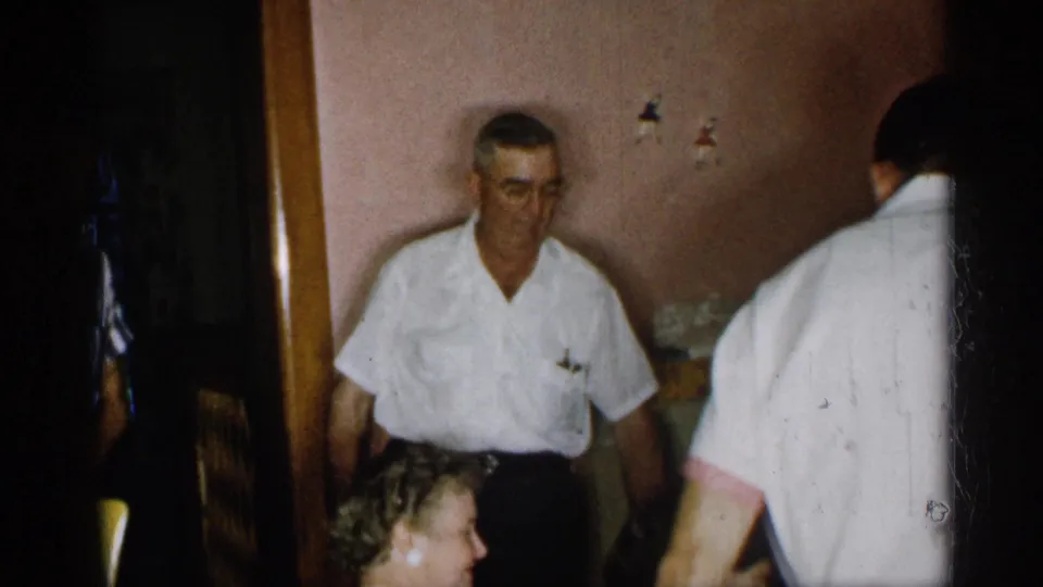 1955 New York home-movie frame showing adults in a private dining room, with a man in a white shirt near seated women.