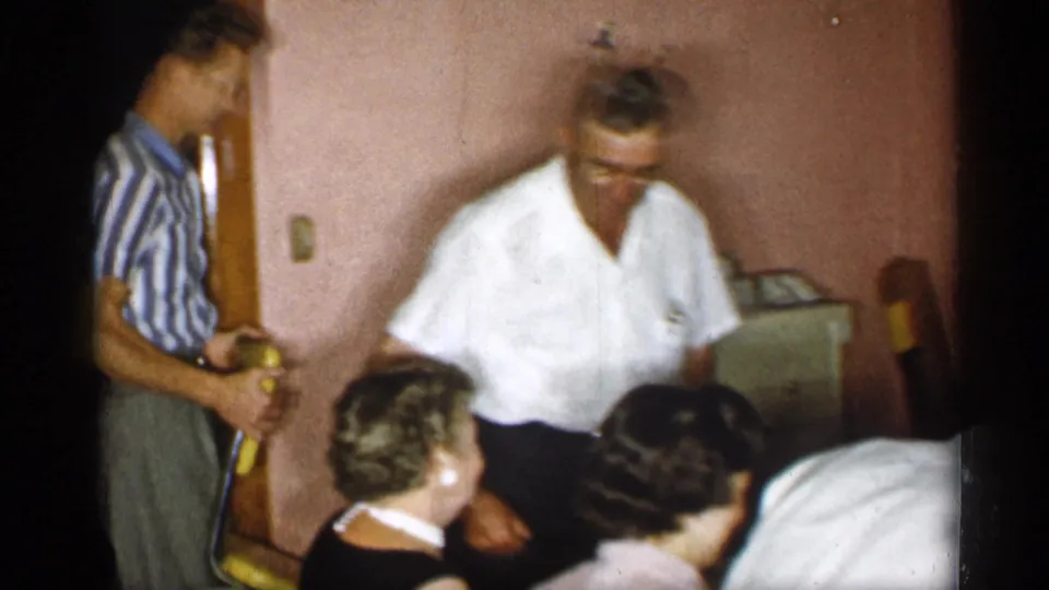 1955 New York home-movie frame showing adults around a dining table, with one man blurred in motion and seated women in the foreground.
