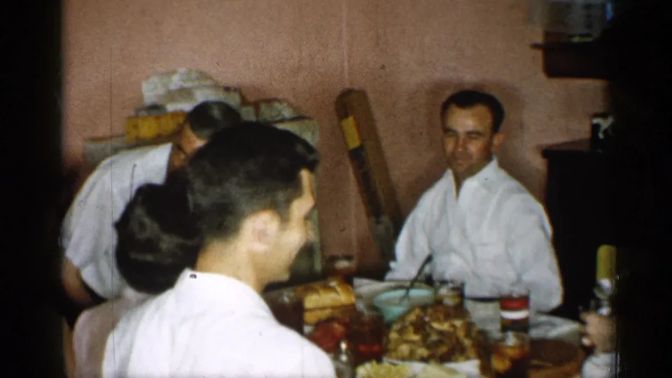 1955 New York family dinner frame with adults seated at a laden table inside a private home.