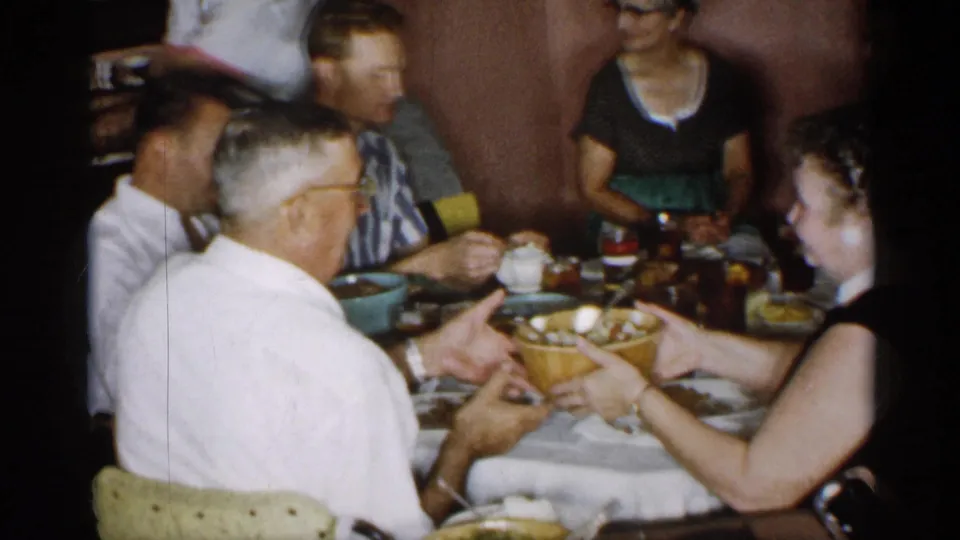 Adults in a 1955 New York family gathering pass a large serving bowl across a crowded dinner table.