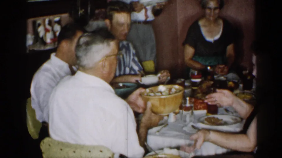 Home-movie view of a mid-1950s New York dinner table with adults seated close together and dishes spread out.