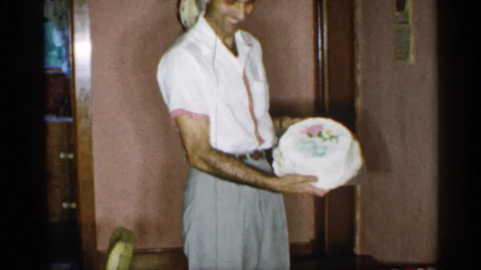 A man in a private New York interior holds a decorated cake, suggesting a 1955 family celebration.