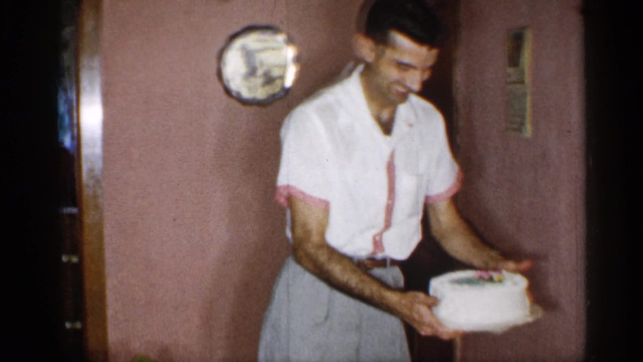 Smiling man holds a small frosted cake in a pink-walled room.