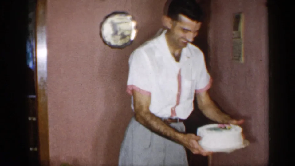 In a 1955 New York home-movie frame, a smiling barefoot man in a white shirt with pink trim carries a frosted cake indoors.