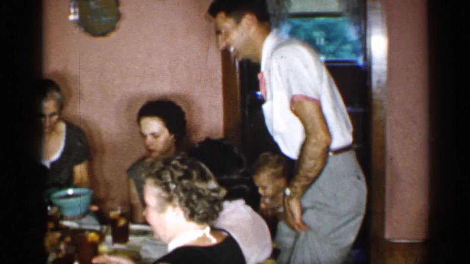 Several family members sit at a dining table in the 1955 New York home while the man stands nearby after bringing the cake.