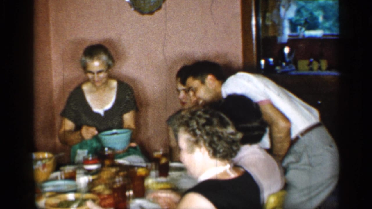 In a 1955 New York home-movie frame, adults gather closely around a small dining table as a woman in glasses holds a blue serving bowl.