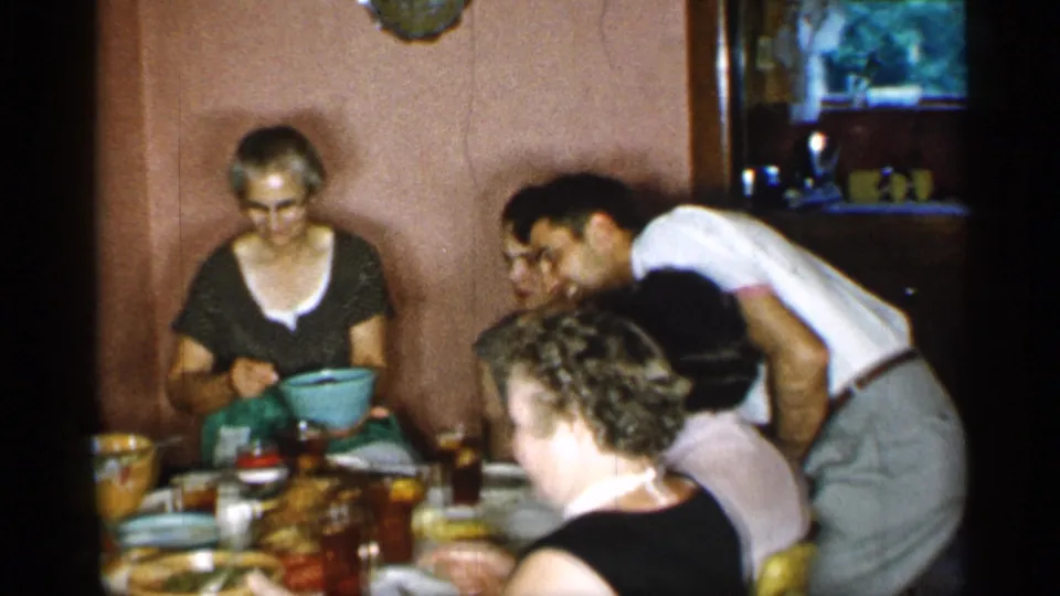 In a 1955 New York home-movie frame, adults gather closely around a small dining table as a woman in glasses holds a blue serving bowl.