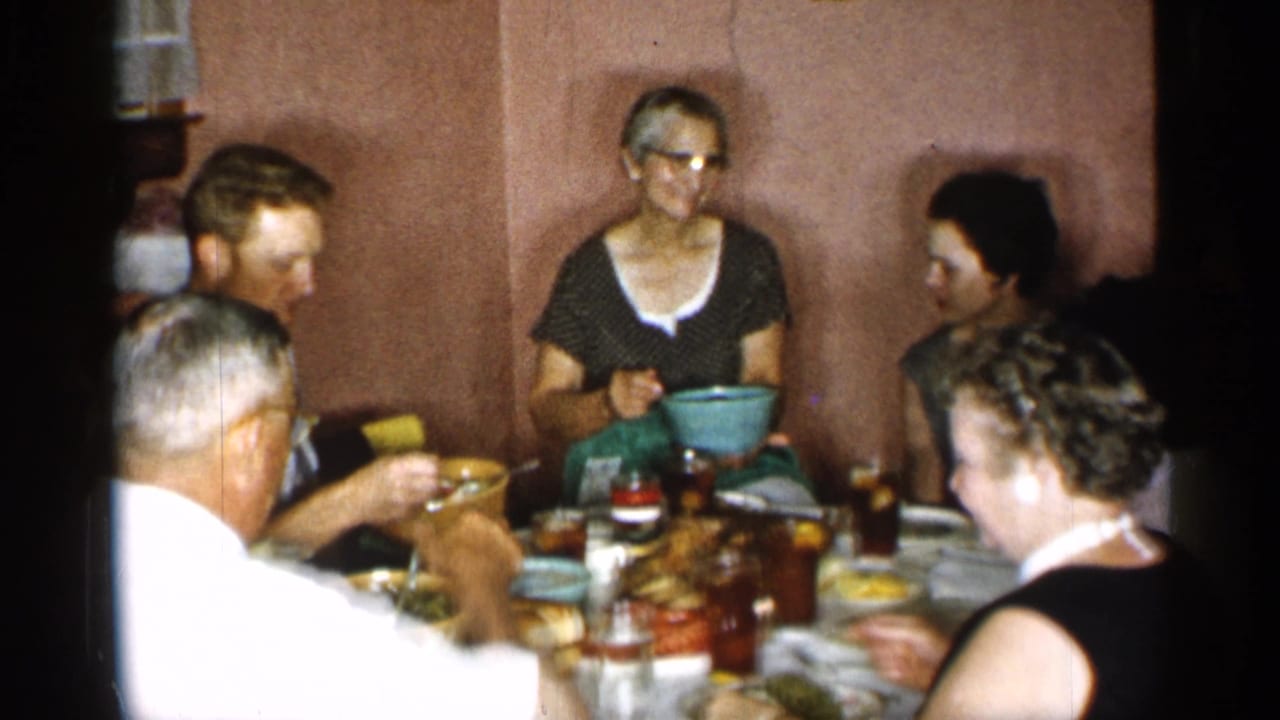 Adults eat at a crowded table while a woman with glasses sits near the back wall.