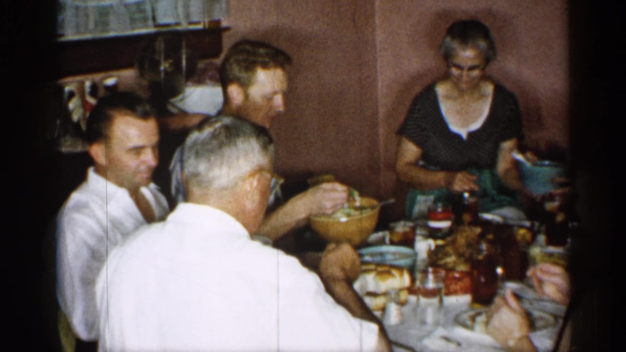 The home-movie frame shows several adults at a small table, with a woman holding a bowl and food, glasses, and serving dishes arranged in front of them.