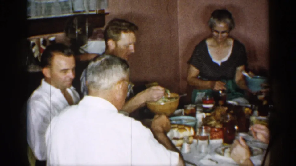 The home-movie frame shows several adults at a small table, with a woman holding a bowl and food, glasses, and serving dishes arranged in front of them.