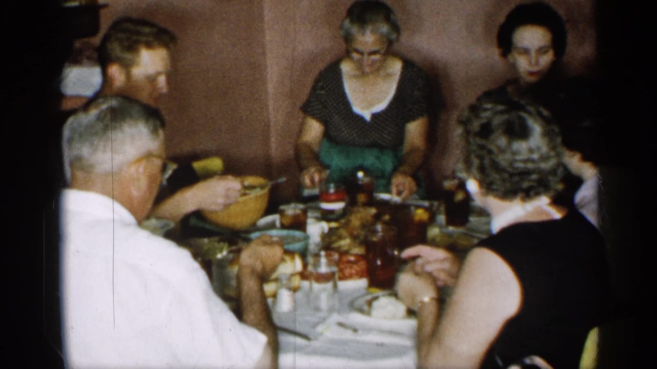 Adults continue a shared meal around a small dining table in the 1955 New York home-movie footage, with bowls, plates, glasses, and close indoor seating visible.